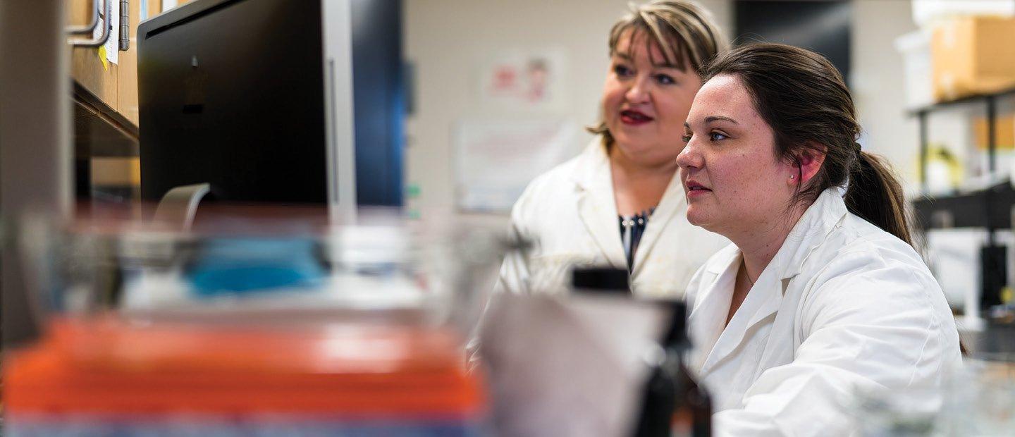 Two women in a white lab coats, looking at a computer screen in a lab.